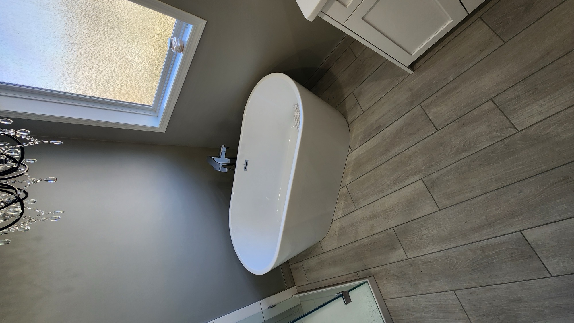 Freestanding soaker tub on wood-look plank tile under a crystal chandelier
